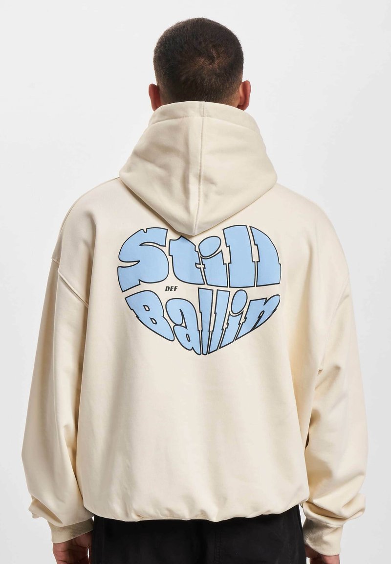 Crème kleurige katoenen hoodie met een ruime capuchon, oversized pasvorm en grote blauwe "Still Ballin"-graphic op de achterkant. Zachte textuur en geribde manchetten.