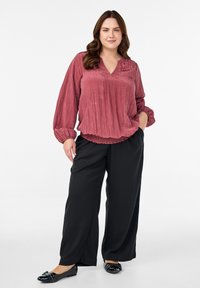 Žena s smeškom i smeškom u smešu koja nosi naboranu ružičastu bluzu s dugim rukavima, crne široke hlače i crne ravne cipele s ukrasima.