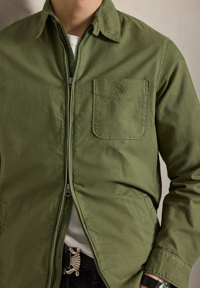 Mann trägt olivgrünes Zip-up-Hemd mit Brusttasche, weißes Unterhemd, schwarzer Gürtel mit Metallschnalle und Uhr am linken Handgelenk.