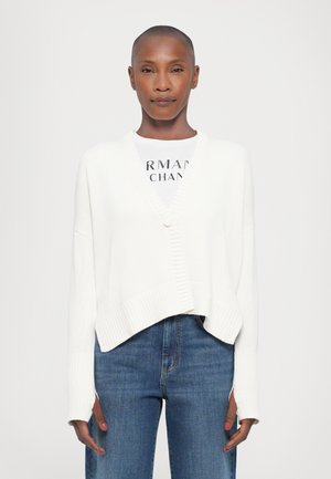 Hvit strikket cardigan med v-hals, ribbestrikkede mansjetter og kant, med en løs passform. Brukt over en hvit grafisk t-skjorte og blå jeans.