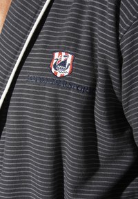 Dunkelgraues Polo mit weißen diagonalen Streifen. Versehen mit einem gewebten Logo-Badge und gesticktem Text auf der Brust. Weicher, strukturierter Stoff.