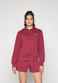 Bordo spalvos džemperis su šortais, pagamintas iš minkštos medžiagos. Džemperis turi garnyrą ir nedidelį spausdintą logotipą krūtinėje. Atsipalaidavęs siluetas.