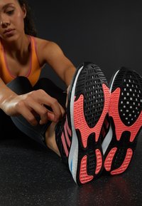 Zwarte sportschoenen met roze accenten en gestructureerde zolen; een persoon die de veters strijkt terwijl hij een zwarte outfit en een oranje top draagt.