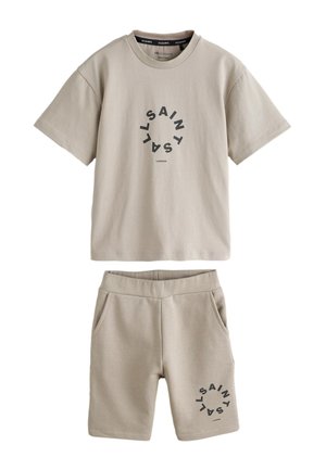 Dreng iført beige matchende t-shirt og shorts med cirkulær sort tekst, hvide sokker og hvide sneakers, står mod en ensfarvet baggrund.