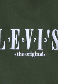 Žalias medvilnės audinys, kuriame užrašyta didelė balta siuvinėta tekstas "LEVI'S" su numeriais 1873 ir žodžiu "originalas" žemiau.