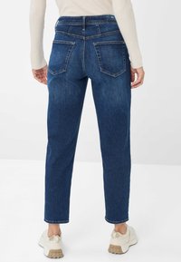 Pantaloni in denim blu scuro con vita alta, vestibilità dritta, due tasche posteriori e dettagli di cucitura chiara, abbinati a sneaker beige.