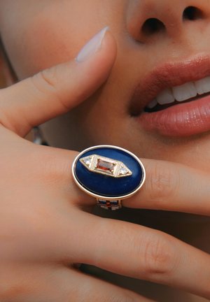 Bague ovale bleue avec une pierre rectangulaire centrale entourée de petits diamants, montée sur un anneau en métal texturé avec des accents colorés.