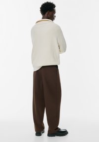 Żółty sweter z prążkowanej wełny z kołnierzykiem, zestawiony z luźnymi brązowymi spodniami i czarnymi butami. Stylizacja ma swobodny, casualowy fason.