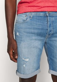 Světle modré denimové kraťasy s viditelným popraskáním a ohrnutými manžetami. Mají klasický 5-kapsový design a stříbrné kovové detaily.