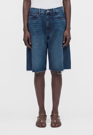 Person iført knælange blå denimshorts med udskæringer i siderne og brune sandaler med tynde stropper og guldornamenter.