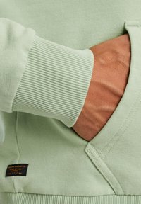 Sweatshirt verde claro con puños acanalados y un bolsillo frontal; presenta un material texturizado y una pequeña etiqueta negra en el dobladillo.