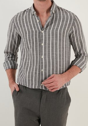 Man draagt een grijs-wit verticaal gestreept overhemd met opgerolde mouwen en donkergrijze broek, hand die de voorkant van het overhemd rechtzet.