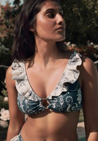 Tumšā teal krāsā ar ziedu rakstu, bikini tops ar dziļu dekoltē, viļņotām malām un centrālo zelta gredzenu. Mīksta auduma tekstūra.