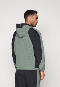 Zelená a čierna nylonová vetrovka s kapucňou, elastickými manžetami a pásom, s kontrastnými lemami a pohodlným strihom.