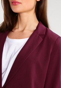 Bordeaux blazer met een smal kraagje, van lichtgewicht stof en een zachte textuur, gelaagd over een witte top. Focus op verfijnde ontwerpdetails.