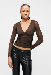 Νεαρό άτομο με πλεξίδες στα μαλλιά φοράει διαφανές καφέ μακρυμάνικο τοπ και μαύρο δερμάτινο παντελόνι, στέκεται με το ένα χέρι στη μέση.