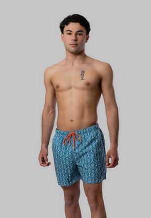 Giovane uomo in piedi senza maglietta, indossa pantaloncini da bagno blu con motivo e coulisse arancione, con tatuaggio nero con caratteri asiatici sul petto.