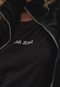 Person med en svart jacka som är öppen och visar en svart skjorta med delvis synlig vit text som står "STAR JEANS."