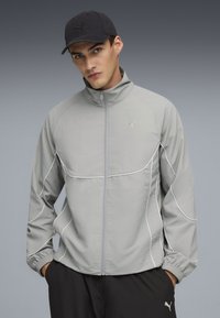 Lys grå zip-jakke med hvide accenter, der har en høj krave og elastiske manchetter. Stoffet ser glat og let ud.