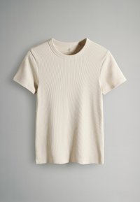 Īsroku krēmkrāsas T-krekls no ribotas auduma ar apaļu izgriezumu un vaļēju piegriezumu, pakārts pret neitrālu fonu.