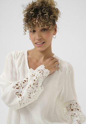 Blusa blanca hecha de una tela ligera con un tejido texturizado. Presenta mangas largas y abullonadas con un intrincado borde de encaje en los puños.