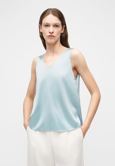 Γυναίκα με καστανά μαλλιά, που φοράει ανοιχτό μπλε σατέν αμάνικο τοπ με λαιμόκοψη σε σχήμα V και λευκό παντελόνι, στέκεται με τα χέρια στις τσέπες μπροστά σε απλό φόντο.