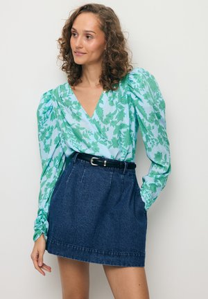 Vrouw met krullend haar, gekleed in een blauw, bloemetjespatroon, blouse met lange mouwen en een donker denim rok met hoge taille en een zwarte riem, staand tegen een witte muur.