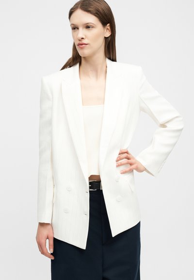 Jeune femme portant un blazer blanc à double boutonnage sur un haut blanc, associée à un pantalon noir, debout avec une main sur la hanche.