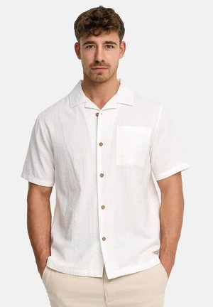 Mand med kort krøllet hår iført en struktureret hvid kortærmet skjorte med knapper og en brystlomme samt beige bukser, hænderne i lommerne.