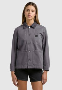 Tânără purtând o jachetă gri cu nasturi și patru buzunare, și pantaloni scurți negri din denim, pe un fundal simplu, deschis la culoare.