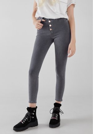 Femme portant un jean skinny gris taille haute avec quatre boutons à l'avant, un shirt blanc à manches courtes et des bottines noires à lacets.