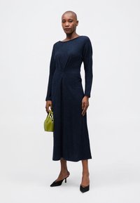 Μακριά μπλουζοφόρεμα σε ναυτικό μπλε με εφαρμοστό κορσέ και φαρδιά φούστα; υφαντό ύφασμα; συνδυασμένο με μια μικρή πράσινη τσάντα και μαύρες ψηλοτάκουνες γόβες.