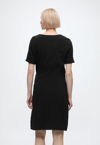 Femme blonde aux cheveux courts portant une robe noire simple mi-longue, debout, dos tourné, sur un fond blanc.