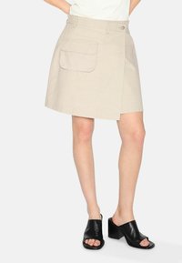 Gonna avvolgente beige con sovrapposizione frontale, una tasca laterale e una texture liscia. Abbinata a mules neri con tacco open-toe.