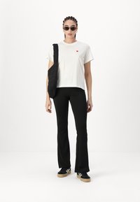 Witte T-shirt met een klein rood hartontwerp, zwarte flared broek, zwarte gewatteerde tas en zwarte sneakers met witte strepen.