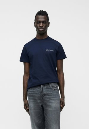 Mees kannab tumesinist Karl Lagerfeld Jeans T-särki ja tuhmiks kulunud halli teksapükse ning seisab ühevärvilise valge tausta ees.