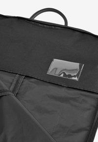 Housse à vêtements noire avec une poignée et une poche d'identification en plastique transparent sur le rabat supérieur, partiellement ouverte pour montrer la doublure intérieure.