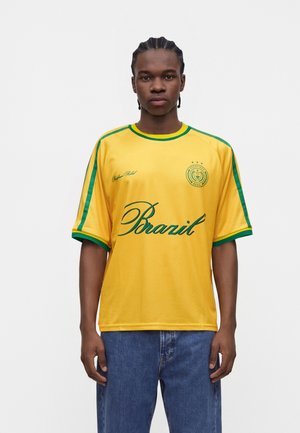 Jeune homme aux cheveux tressés portant un maillot de sport jaune du Brésil avec une bordure verte et un jean bleu, face à un fond uni.