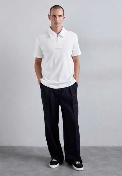 Un homme adossé à un mur gris, portant un polo blanc à manches courtes avec fermeture éclair, un pantalon noir à jambes larges et des baskets noires avec des lacets blancs.
