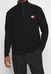 Suéter de media cremallera negro con canalé y un alto cuello, que presenta un pequeño parche de logo en rojo, blanco y azul en el lado izquierdo del pecho.