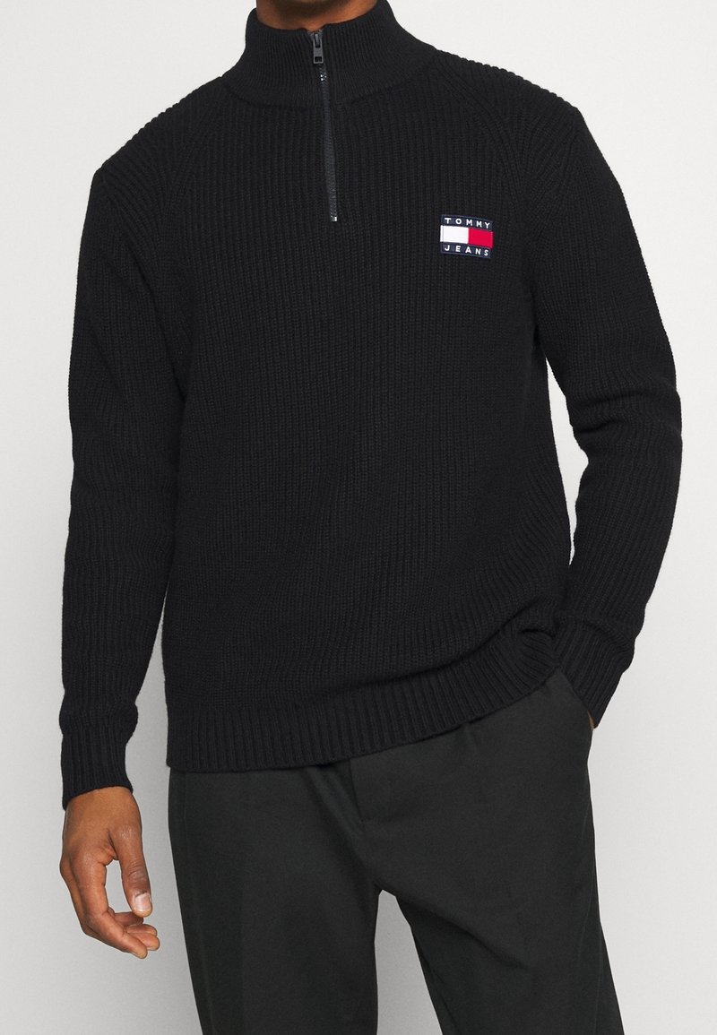 Suéter de media cremallera negro con canalé y un alto cuello, que presenta un pequeño parche de logo en rojo, blanco y azul en el lado izquierdo del pecho.