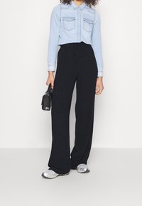 Kvinna i ljusblå knäppt skjorta, lösa svarta byxor, silversneakers, som håller en liten svart handväska vid sin sida.