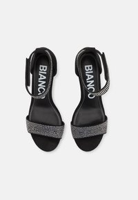 Sandali neri in suede con una fascia decorata da strass. Presentano un tacco basso e una strap regolabile alla caviglia. La suola è liscia e scura.