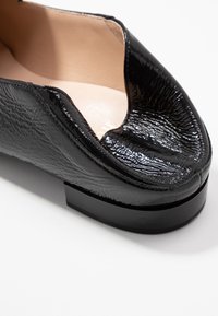 Chaussure en cuir noir avec une finition brillante et texturée, design à bout ouvert et un talon bas et arrondi. Doublure intérieure lisse.