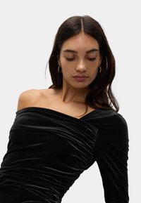 Czarna aksamitna sukienka z asymetrycznym, jedno-ramiennym fasonem, z marszczonymi detalami i gładką fakturą. Złote kolczyki w kształcie koła dopełniają stylizację.