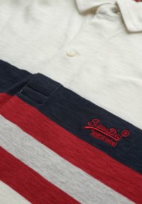 Hvid polo shirt med marineblå og røde vandrette striber, med en knaplukning og broderet "SuperDry" logo i rød. Blød tekstur.