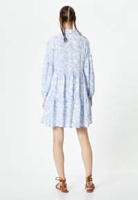 Vestido floral azul y blanco con mangas largas abullonadas, cintura fruncida y falda en capas. Combinado con sandalias marrones de tiras, textura de tela suave.