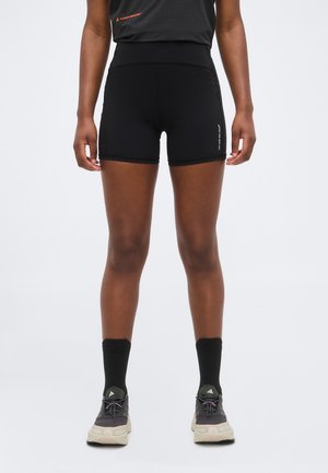 Person iført svarte treningsshorts, svarte sokker og grå Adidas-sneakers, stående foran en ensfarget lys bakgrunn.