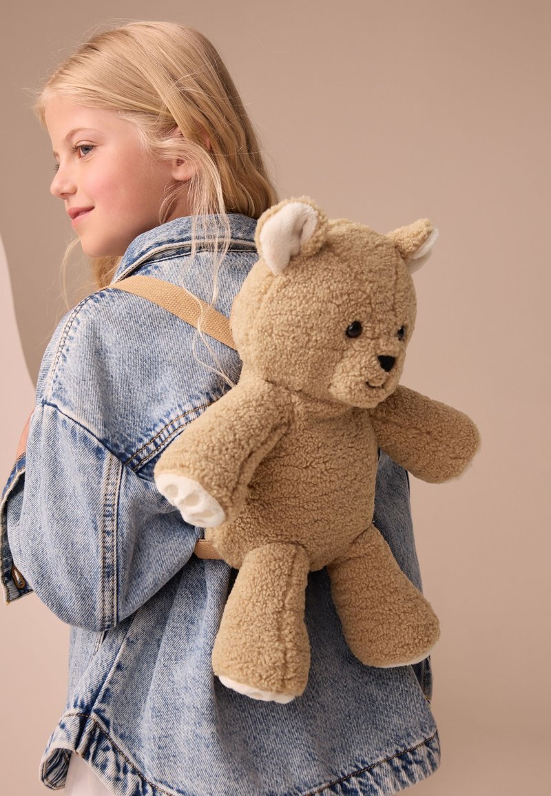 Ung pige med blondt hår iført en denimjakke og med en beige bamserygsæk på ryggen mod en neutral baggrund.