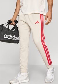 Jasnobeżowe spodnie dresowe z trzema czerwonymi pionowymi paskami, z haftowanym czerwonym logo Adidas na udzie. Towarzyszy im czarna torba.